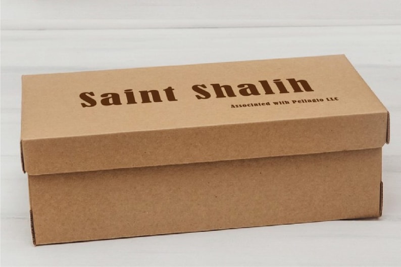 Puede incluir: Caja rectangular de cart&oacute;n marr&oacute;n claro con tapa. En la parte superior, en color marr&oacute;n oscuro, se lee "Saint Shalih". Debajo del texto est&aacute; escrito "Associated with Pellagio LLC".