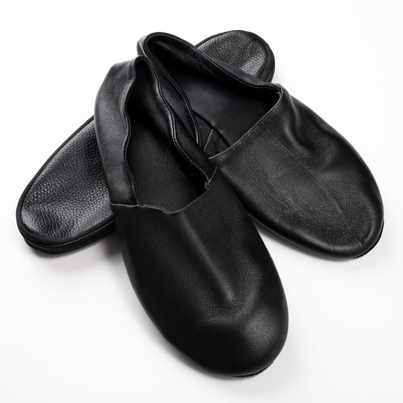 May include: Black leather ballet slippers with a soft, rounded toe. The slippers are shown with a matching black zippered carrying case. The leather has a slight sheen, and the slippers appear to be unworn.
