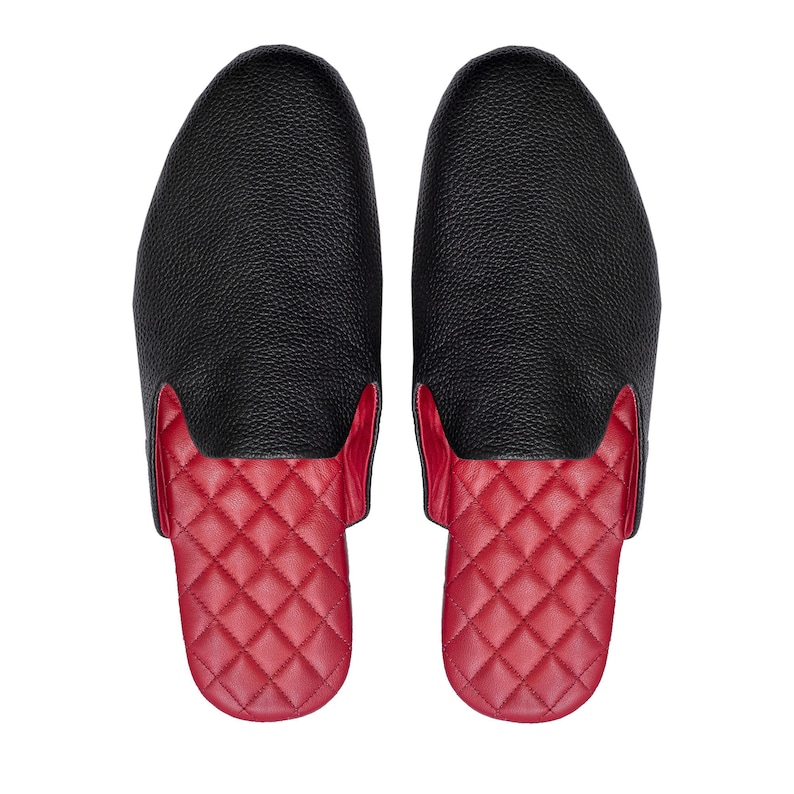 May include: A pair of black leather slippers with a textured finish. The toe and upper portion are black, while the footbed is red with a quilted diamond pattern. The slippers are designed for indoor use.