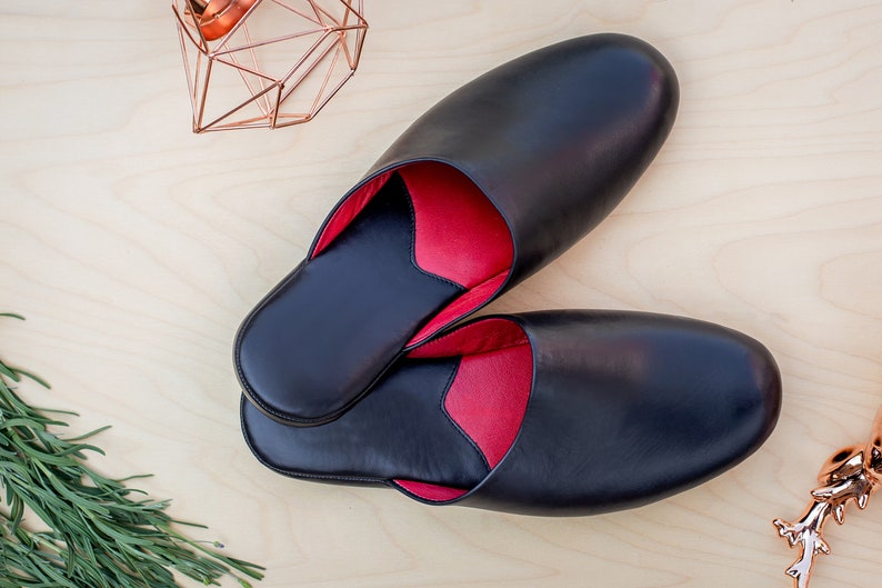 Puede incluir: Un par de zapatillas de cuero negro con interior rojo. Las zapatillas son de forma ovalada y parecen nuevas. La imagen est&aacute; tomada desde arriba, mostrando las zapatillas sobre una superficie de madera clara.