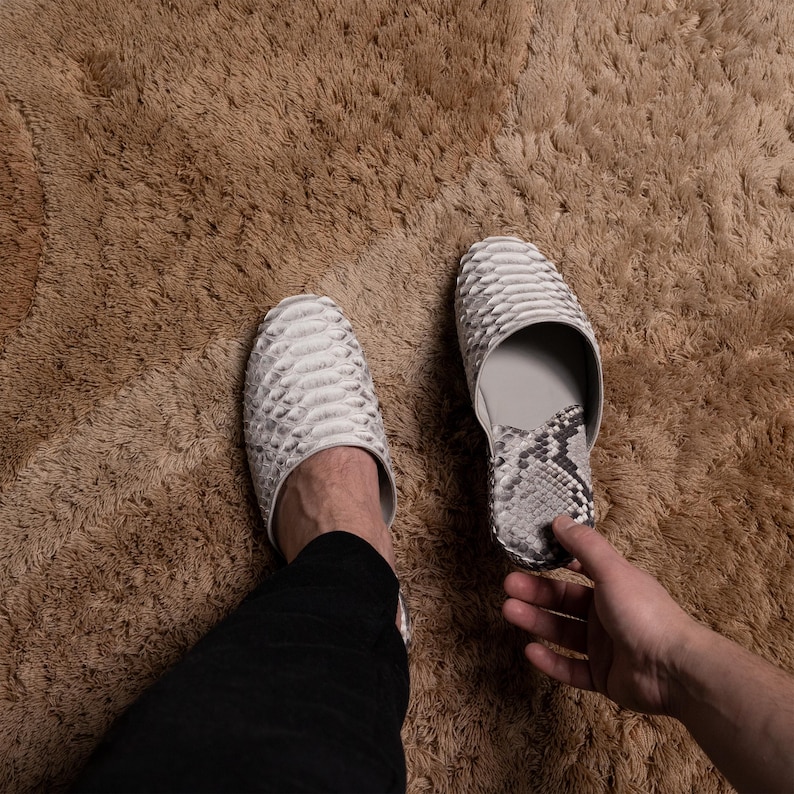 May include: A pair of white snake skin slippers with a textured pattern. The slippers are worn on a person's feet.