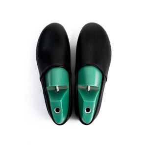 May include: A pair of black leather shoe covers with green shoe trees inside. The shoe trees are a bright green color and have a metal piece at the top. The covers are smooth and have a rounded toe.