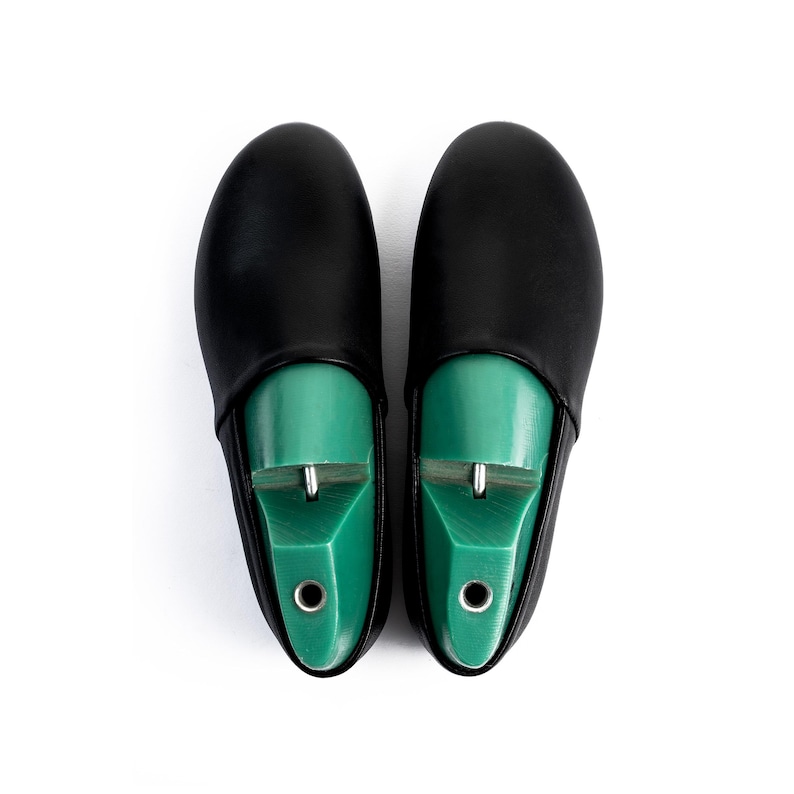 May include: A pair of black leather shoe covers with green shoe trees inside. The shoe trees are a bright green color and have a metal piece at the top. The covers are smooth and have a rounded toe.