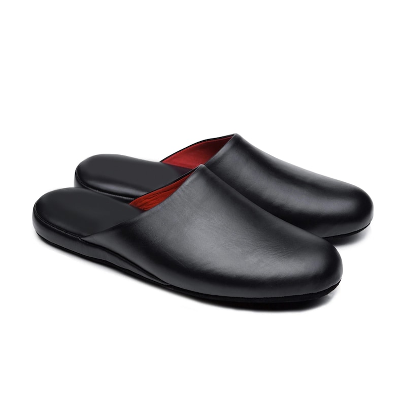 Puede incluir: Un par de zapatillas negras de cuero con una textura suave y forro interior rojo. Estas zapatillas de interior se presentan sobre un fondo blanco.