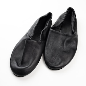 May include: A pair of black leather slippers, designed with a rounded toe and a soft, flexible upper. The slippers are shown on a white background, highlighting their simple, classic design. These slippers are suitable for indoor wear.