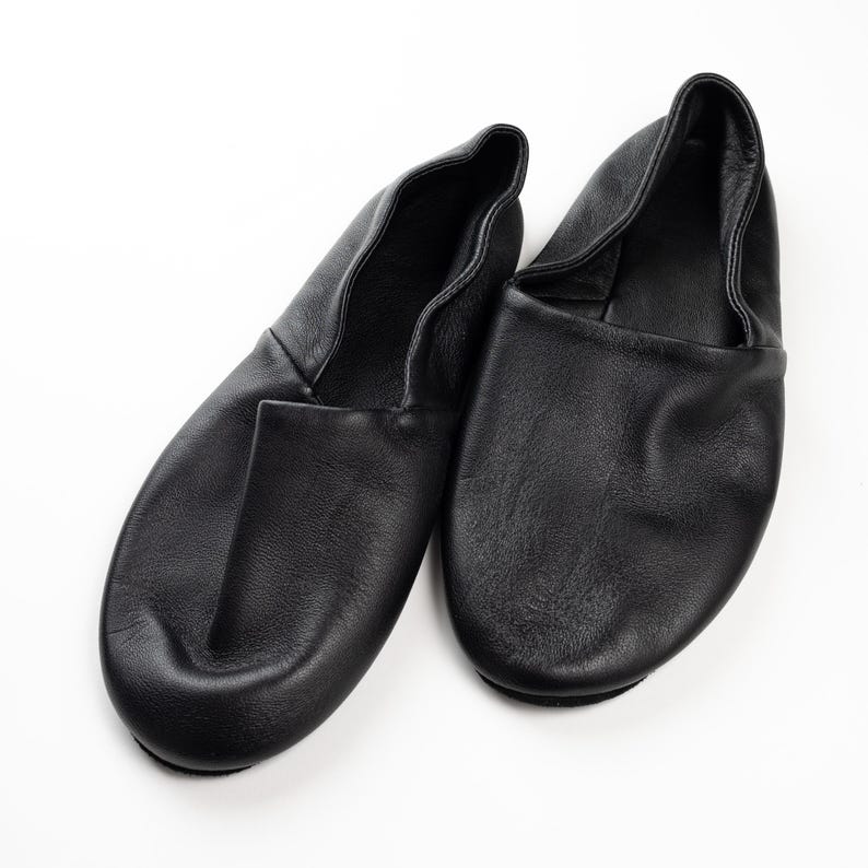 May include: A pair of black leather slippers, designed with a rounded toe and a soft, flexible upper. The slippers are shown on a white background, highlighting their simple, classic design. These slippers are suitable for indoor wear.