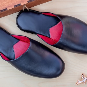Puede incluir: Un par de zapatillas de cuero negro con interior rojo, presentadas sobre una superficie de madera clara. Las zapatillas tienen un dise&ntilde;o cl&aacute;sico, con punta redondeada y estilo sin cordones. La imagen est&aacute; bien iluminada, mostrando la textura y calidad del cuero.