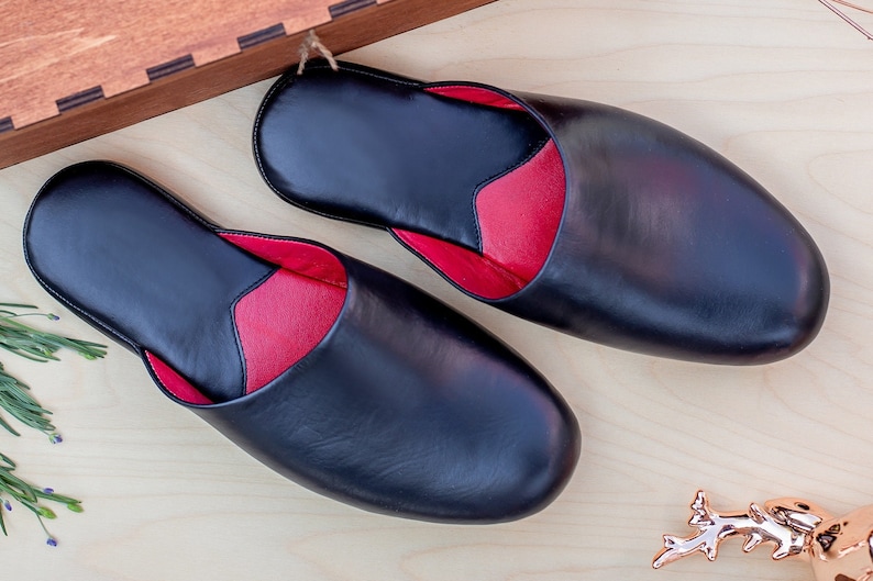 Puede incluir: Un par de zapatillas de cuero negro con interior rojo, presentadas sobre una superficie de madera clara. Las zapatillas tienen un dise&ntilde;o cl&aacute;sico, con punta redondeada y estilo sin cordones. La imagen est&aacute; bien iluminada, mostrando la textura y calidad del cuero.