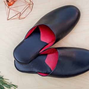Puede incluir: Un par de zapatillas de cuero negro con interior rojo. Las zapatillas, de forma ovalada, parecen nuevas. La foto est&aacute; tomada desde arriba, con formas geom&eacute;tricas cobrizas y vegetaci&oacute;n en el fondo. Ideales para uso en interiores.