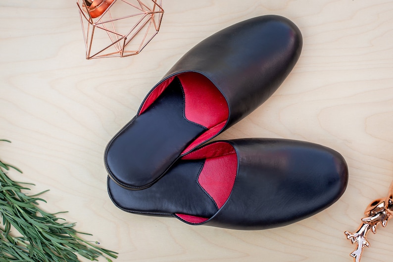 Puede incluir: Un par de zapatillas de cuero negro con interior rojo. Las zapatillas, de forma ovalada, parecen nuevas. La foto est&aacute; tomada desde arriba, con formas geom&eacute;tricas cobrizas y vegetaci&oacute;n en el fondo. Ideales para uso en interiores.