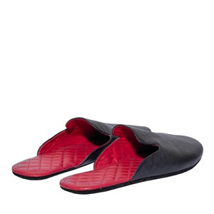 May include: A pair of black leather slippers with a red quilted interior. The slippers have a slip-on design and a rounded toe. The exterior leather has a textured appearance, and the interior is a vibrant red color.