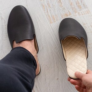 May include: A pair of black leather slippers with a white quilted lining. One slipper is on a person's foot, and the other is being held in a hand.