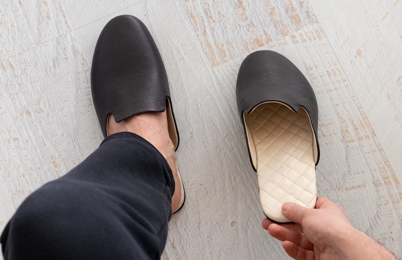 May include: A pair of black leather slippers with a white quilted lining. One slipper is on a person's foot, and the other is being held in a hand.