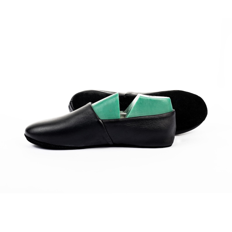 May include: Black leather slip-on shoes with a rounded toe. The shoes have a simple design and are shown with a green shoe tree inside. The second shoe is visible in the background, with the sole facing up.