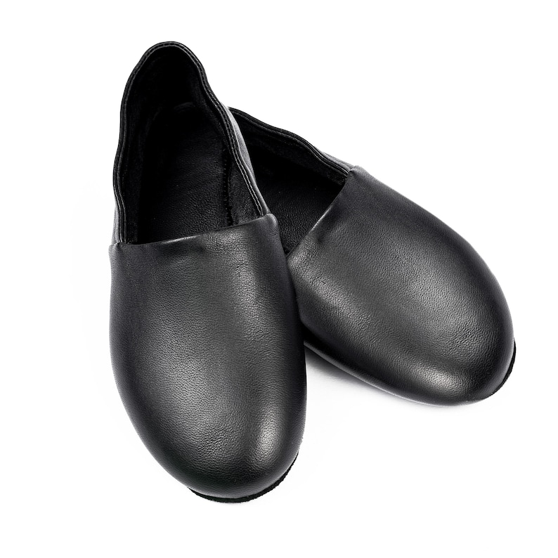 May include: A pair of black leather ballet slippers. The shoes are smooth and have a rounded toe. The slippers are designed to be worn by dancers. The shoes are shown on a white background.