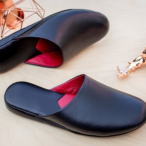 Puede incluir: Zapatillas de cuero negro con interior rojo, expuestas sobre una superficie de madera clara. Las zapatillas son abiertas por detr&aacute;s y tienen una puntera redondeada. Tambi&eacute;n hay formas geom&eacute;tricas cobrizas y una figura de escarabajo.