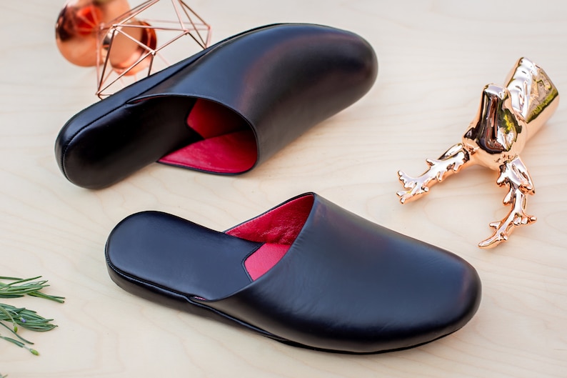 Puede incluir: Zapatillas de cuero negro con interior rojo, expuestas sobre una superficie de madera clara. Las zapatillas son abiertas por detr&aacute;s y tienen una puntera redondeada. Tambi&eacute;n hay formas geom&eacute;tricas cobrizas y una figura de escarabajo.