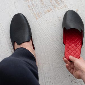 May include: Black leather slipper with a red quilted interior. The slipper has a rounded toe and a low-cut design. The image shows one slipper on a foot and another held by a hand, showcasing the contrasting colors and textures.