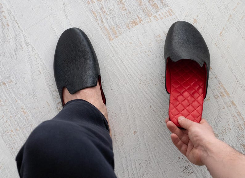 May include: Black leather slipper with a red quilted interior. The slipper has a rounded toe and a low-cut design. The image shows one slipper on a foot and another held by a hand, showcasing the contrasting colors and textures.