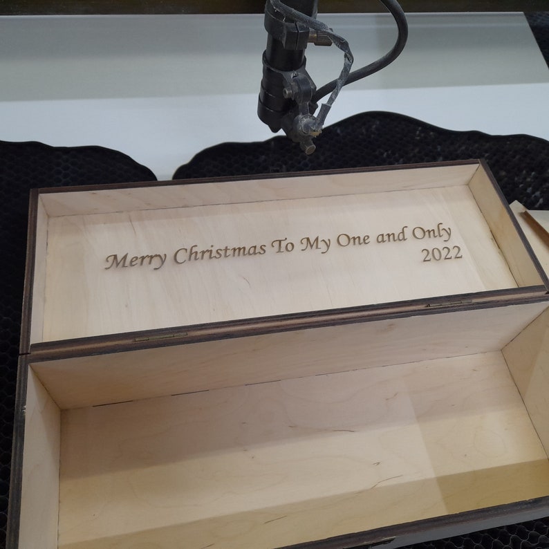 May include: A wooden box with a hinged lid. The lid is open and the inside of the box is visible. The lid is engraved with the text "Merry Christmas To My One and Only 2022".