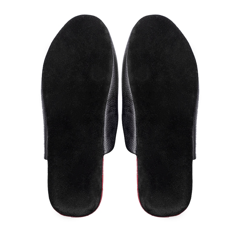 May include: A pair of black leather slippers, viewed from the bottom, showcasing a textured surface. The interior features a soft, dark material with a red accent along the edges. The design is simple and elegant.