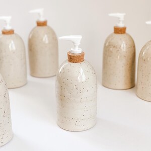Six wheel-thrown, speckled stoneware soap pumps with cork sleeves and white plastic pumps in and our of the frame with one in the centre in focus on white background