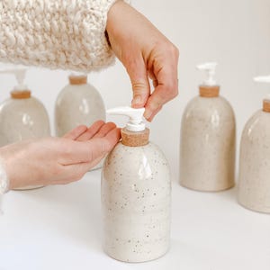 Five wheel-thrown, speckled stoneware soap pumps with cork sleeves and white plastic pumps in and our of the frame with one in the centre in focus with a white woman's hands in a cream knit sweater, dispensing soap into hand on white background