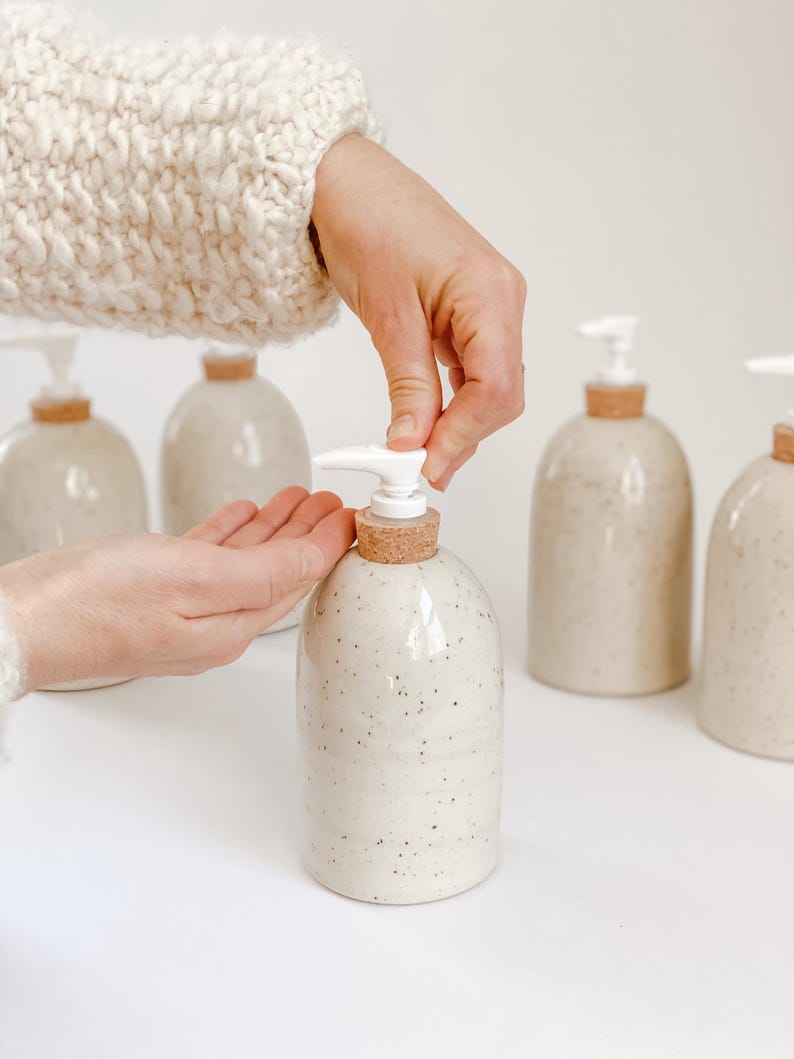 Five wheel-thrown, speckled stoneware soap pumps with cork sleeves and white plastic pumps in and our of the frame with one in the centre in focus with a white woman's hands in a cream knit sweater, dispensing soap into hand on white background