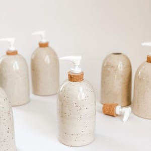 Six wheel-thrown, speckled stoneware soap pumps with cork sleeves and white plastic pumps in and our of the frame with one in the centre in focus and one in the background with the cork and pump removed on white background