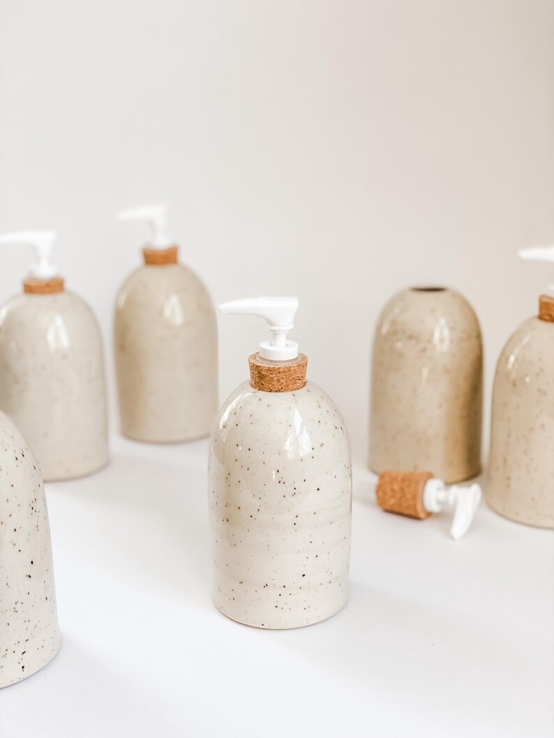 Six wheel-thrown, speckled stoneware soap pumps with cork sleeves and white plastic pumps in and our of the frame with one in the centre in focus and one in the background with the cork and pump removed on white background