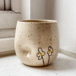 May include: A speckled, cream-coloured ceramic tumbler with a rounded shape. Two simple yellow and black flower illustrations are hand-painted on the side. The cup has a unique, slightly indented design feature.