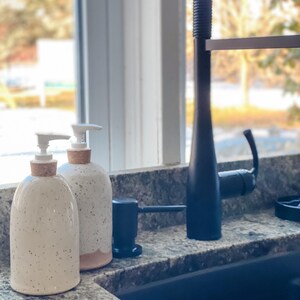 Two wheel-thrown speckled stoneware soap pumps with cork sleeves and white pumps sitting on a granite counter top beside a modern kitchen sink with a window behind