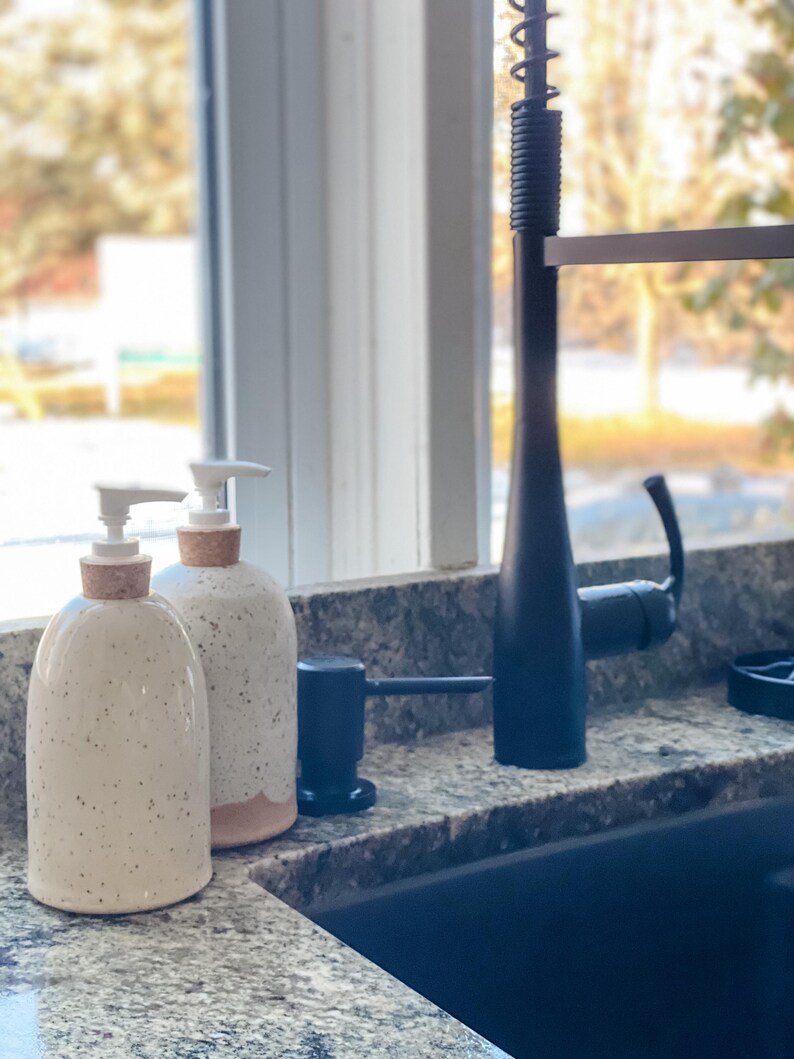 Two wheel-thrown speckled stoneware soap pumps with cork sleeves and white pumps sitting on a granite counter top beside a modern kitchen sink with a window behind