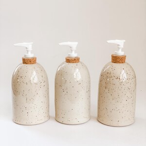 Three wheel-thrown, speckled stoneware soap pumps with cork sleeves and white plastic pumps in a row on white background