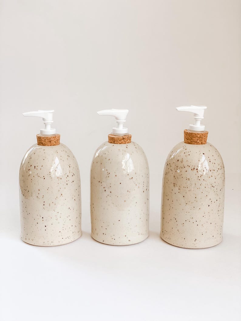 Three wheel-thrown, speckled stoneware soap pumps with cork sleeves and white plastic pumps in a row on white background