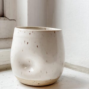 May include: A speckled, off-white ceramic tumbler with a rounded body and a natural, unglazed base. The cup features a subtle indentation on one side and small brown speckles throughout. The interior is smooth and white.
