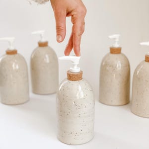 Five wheel-thrown, speckled stoneware soap pumps with cork sleeves and white plastic pumps in and our of the frame with one in the centre in focus with a white woman's hand in a cream knit sweater, touching the top on white background