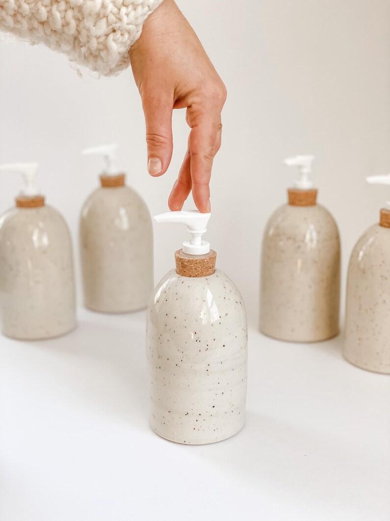 Five wheel-thrown, speckled stoneware soap pumps with cork sleeves and white plastic pumps in and our of the frame with one in the centre in focus with a white woman's hand in a cream knit sweater, touching the top on white background
