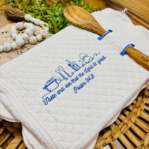 Könnte beinhalten: Weißes, gestepptes Küchentuch mit blau gesticktem Text und Besteckdesign. Der Text lautet „Schmecket und sehet, wie freundlich der HERR ist, Psalm 34,8“. Zwei Holzlöffel mit blauen Akzenten sind sichtbar.
