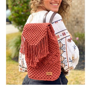 Könnte beinhalten: Ein rostfarbener Makramee-Rucksack mit Klappe und Fransen. Die Tasche hat braune Lederriemen und ein rechteckiges Etikett mit Text. Die Tasche ist aus gewebter Baumwolle, ein stilvolles Accessoire.