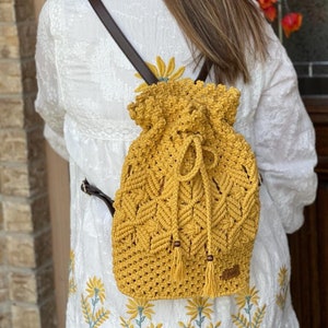 May include: Mustard yellow macrame backpack with brown leather straps. The drawstring closure backpack features a textured, woven design with tassels and wooden beads. The bag is worn over a white embroidered top.