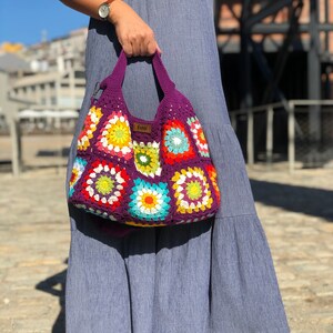 May include: A handmade crochet tote bag in a vibrant patchwork design. The bag features a deep purple base with colorful floral squares in shades of red, yellow, green, and blue. The bag has a matching purple handle and a small label that says "ROME".