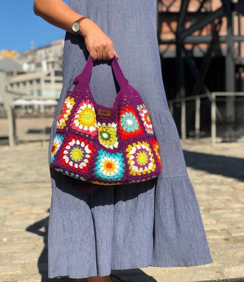 May include: A handmade crochet tote bag in a vibrant patchwork design. The bag features a deep purple base with colorful floral squares in shades of red, yellow, green, and blue. The bag has a matching purple handle and a small label that says "ROME".