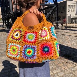 May include: A mustard yellow crochet tote bag with colorful granny squares. The bag features a rectangular shape with a wide strap and a small brown tag that reads "RoMe". The bag is held over the shoulder.