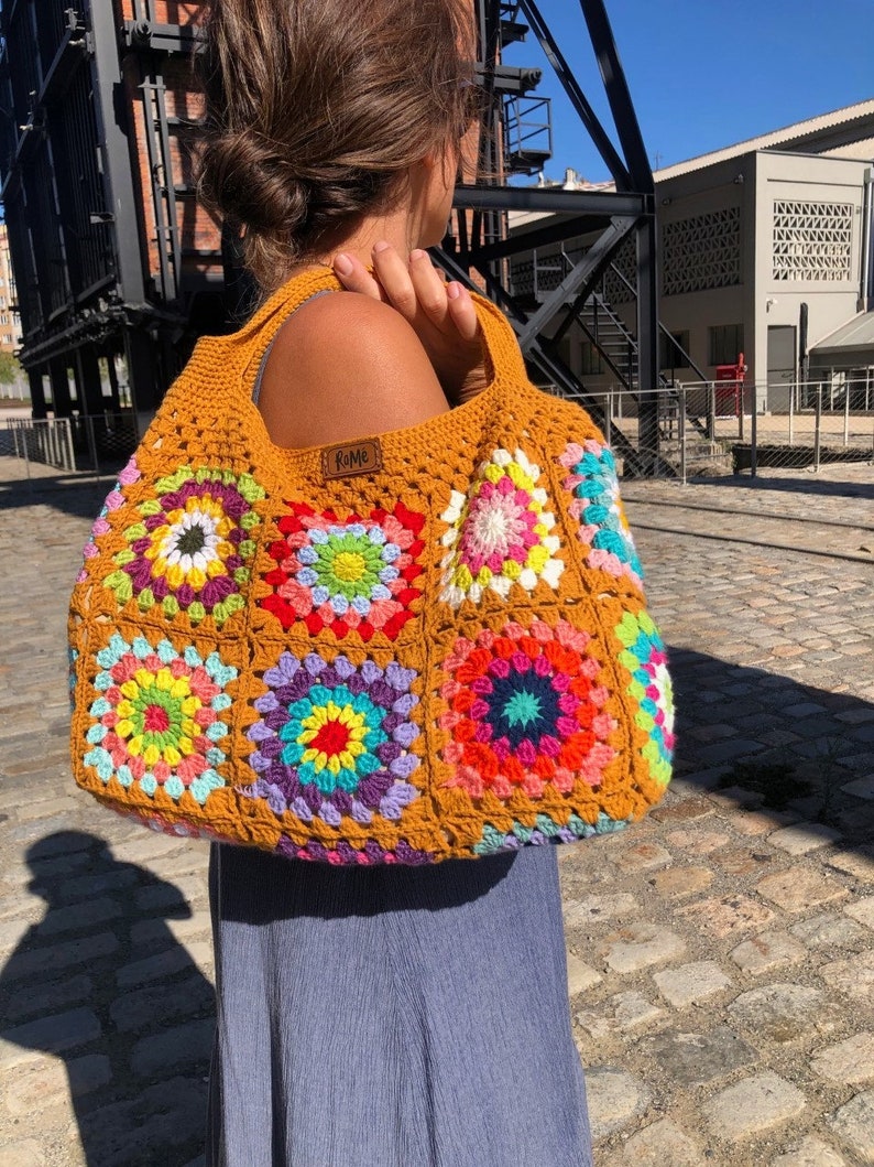 May include: A mustard yellow crochet tote bag with colorful granny squares. The bag features a rectangular shape with a wide strap and a small brown tag that reads "RoMe". The bag is held over the shoulder.