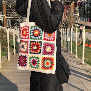May include: A white crocheted tote bag with colorful granny square designs. The bag has a long strap and is being held by a person wearing a black jacket.