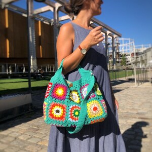 May include: A colorful, handmade crochet bag with a turquoise green strap and body. The bag features square patterns in vibrant colors like red, yellow, orange, and green. The bag is carried over the shoulder.
