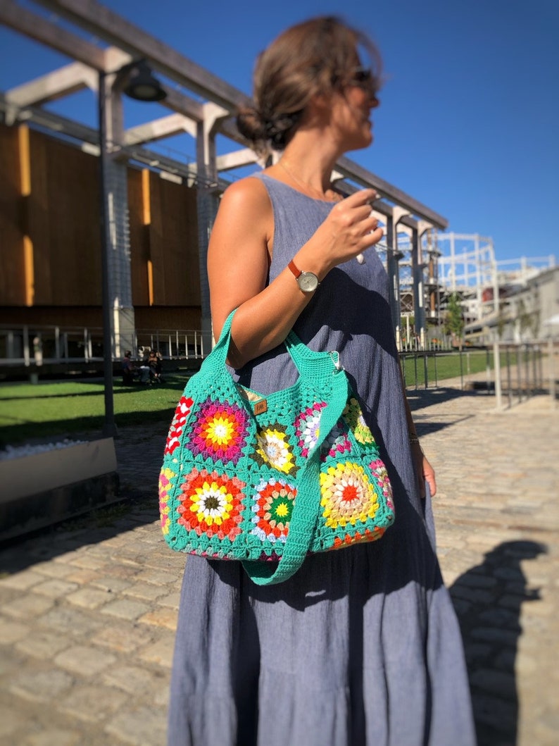 May include: A colorful, handmade crochet bag with a turquoise green strap and body. The bag features square patterns in vibrant colors like red, yellow, orange, and green. The bag is carried over the shoulder.