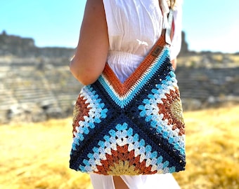 May include: A crocheted tote bag with a geometric pattern in shades of blue, white, brown, and orange. The bag is worn over the shoulder of a person.