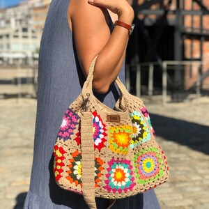 May include: A colorful, crocheted tote bag with a patchwork design. The bag features squares of various colors, including pink, yellow, orange, and blue, with a beige strap and handle. The bag has a label that reads "Rome".